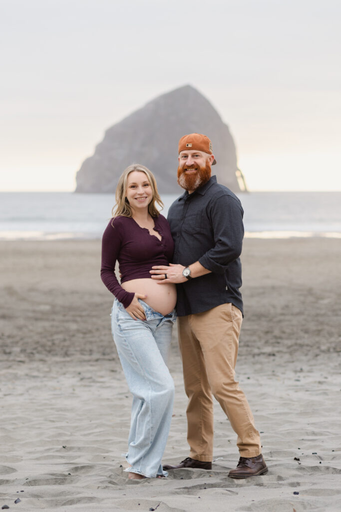 Husband and wife stand in front of Haystack rock at Cape Kiwanda for maternity portrait on the beach.