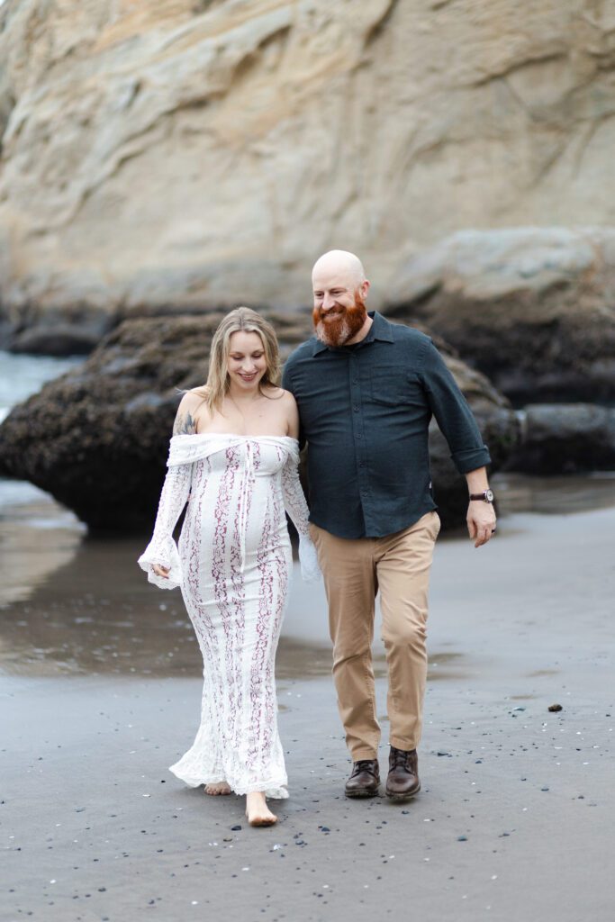 Couple walks along the beach holding hands and smiling for their maternity photos, Portland.