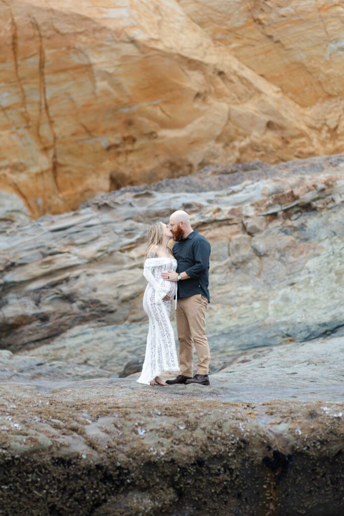 Couple share a kiss on top of a rocky coast in Cape Kiwanda for their maternity portraits.