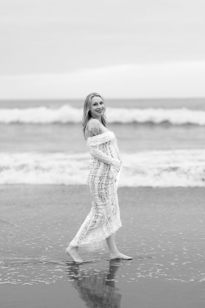 Pregnant mother walks on the waters edges with ocean waves behind her for this black and white portrait.