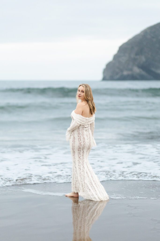 Pregnant mom in a flowy dress walks out into the ocean waves while looking toward the camera for maternity beach session.
