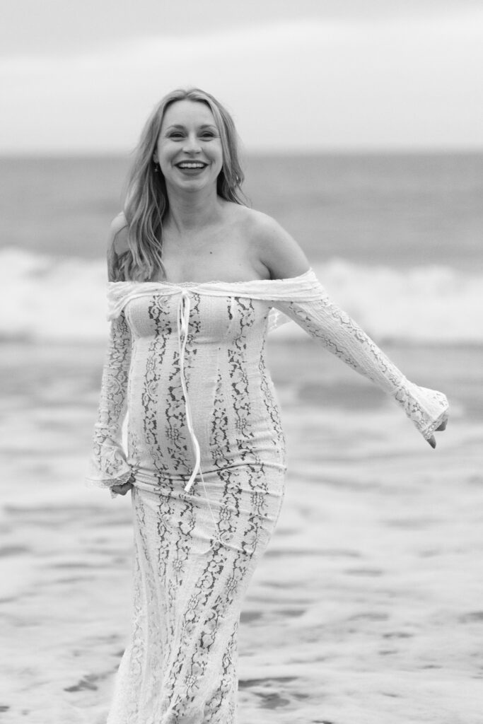 Pregnant mother wearing white dress laughs while walking in the waves during her maternity session.