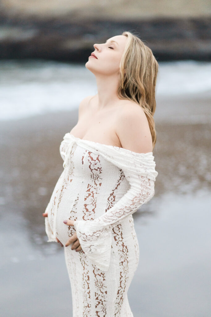 Pregnant mother by the ocean, closes her eyes and embraces her growing baby inside her belly while lifting her chin to the sky.