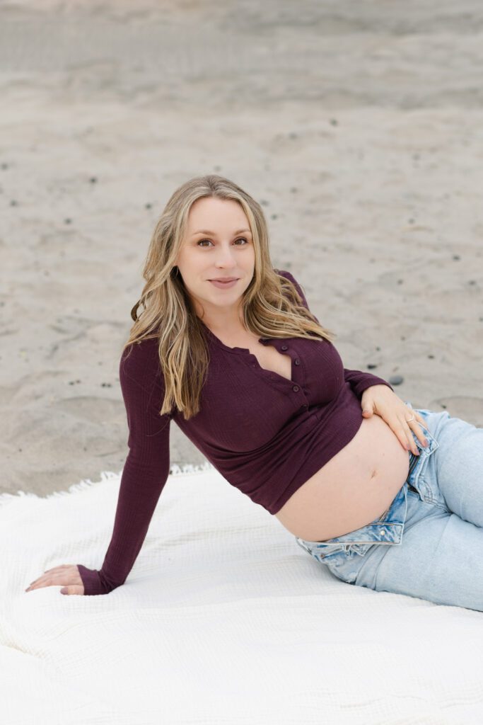 Pregnant mother places her hand on her growing baby bell while sitting on a blanket at the beach for maternity photos, Portland