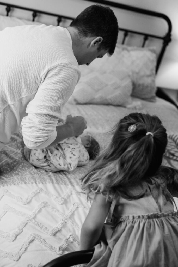 Dad swaddles newborn baby with big sister watching for this candid moment.