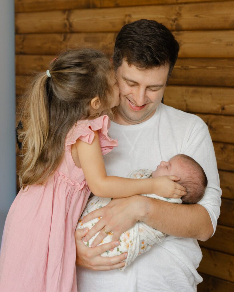 Proud father of two girls, cradles his newborn daughter while his other daughter gives him a kiss on the cheek for family portraits.