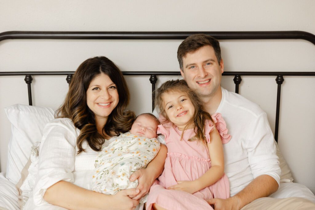 Family portrait by Newborn photographer Portland sitting on a bed for an in home family session with parents holding baby and big sister.