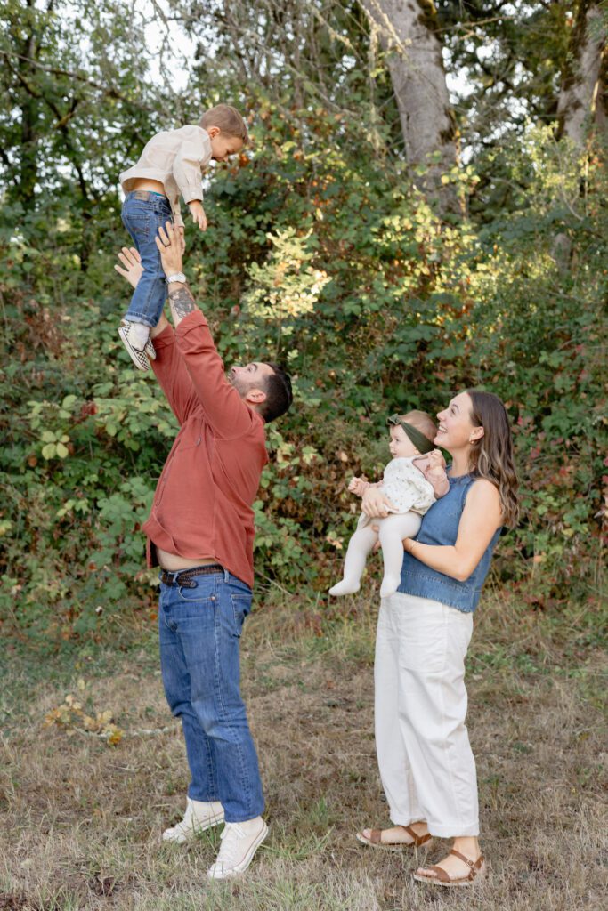 Family plays together at Portland lifestyle family photographer's photo session. The dad throws his little boy in the air and the mom and little sister smile at them.