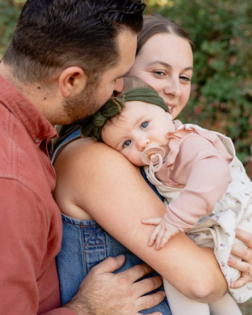 A tender moment with couple and their baby. Young baby looks at camera while hugging her mom, while her dad kisses her forehead.