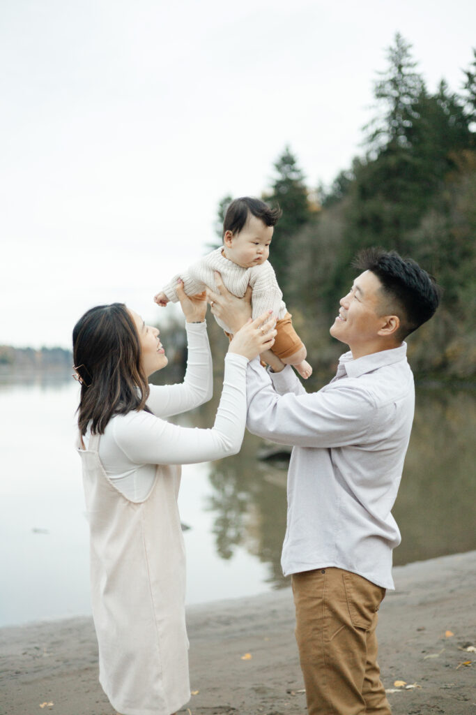 Portland lifestyle family photographer prompts couple to lift their young child in the air with lake and trees behind them.