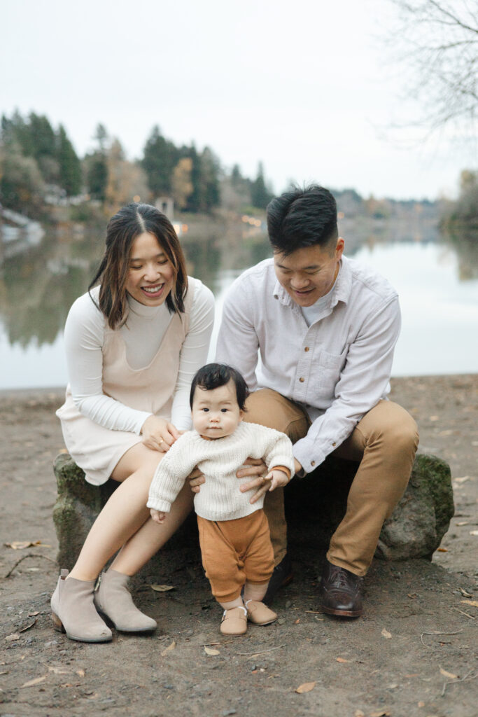 Couple sits on rock near river smiling at their young son who is looking up at camera.