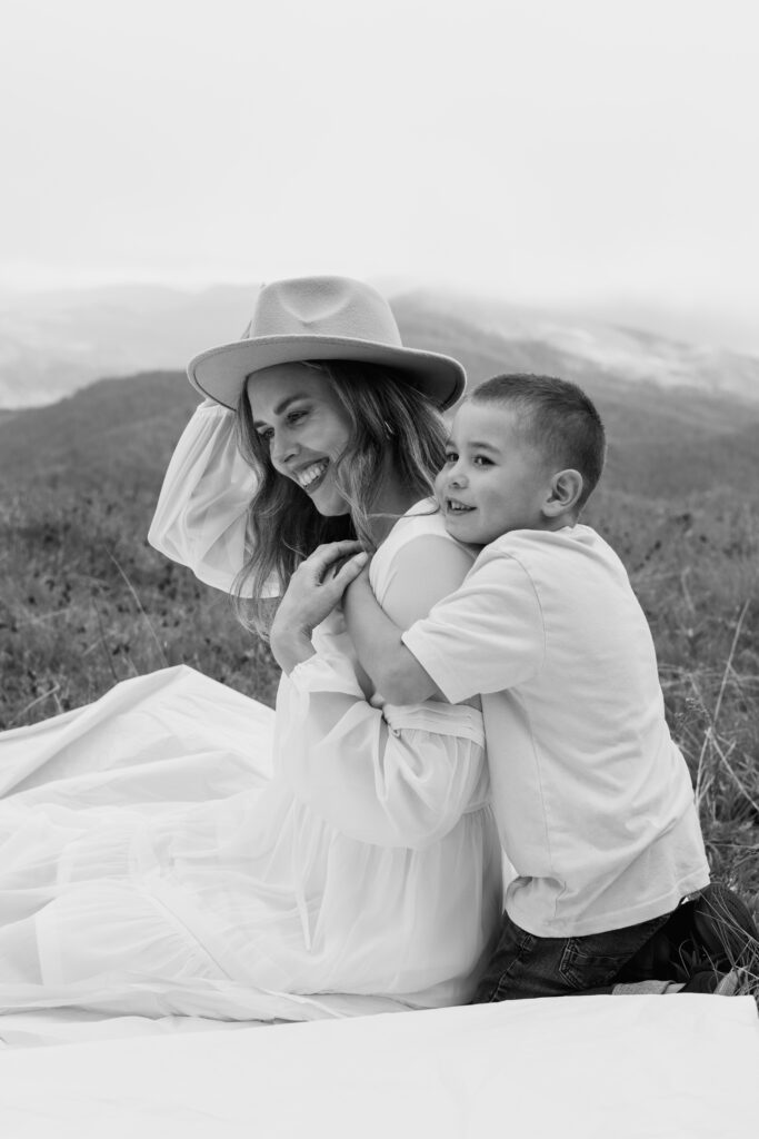 Black and white photo of little boy who is hugging his mom from behind as she sits on blanket, during this mountain top outdoor session at Mary's Peak, Oregon.
